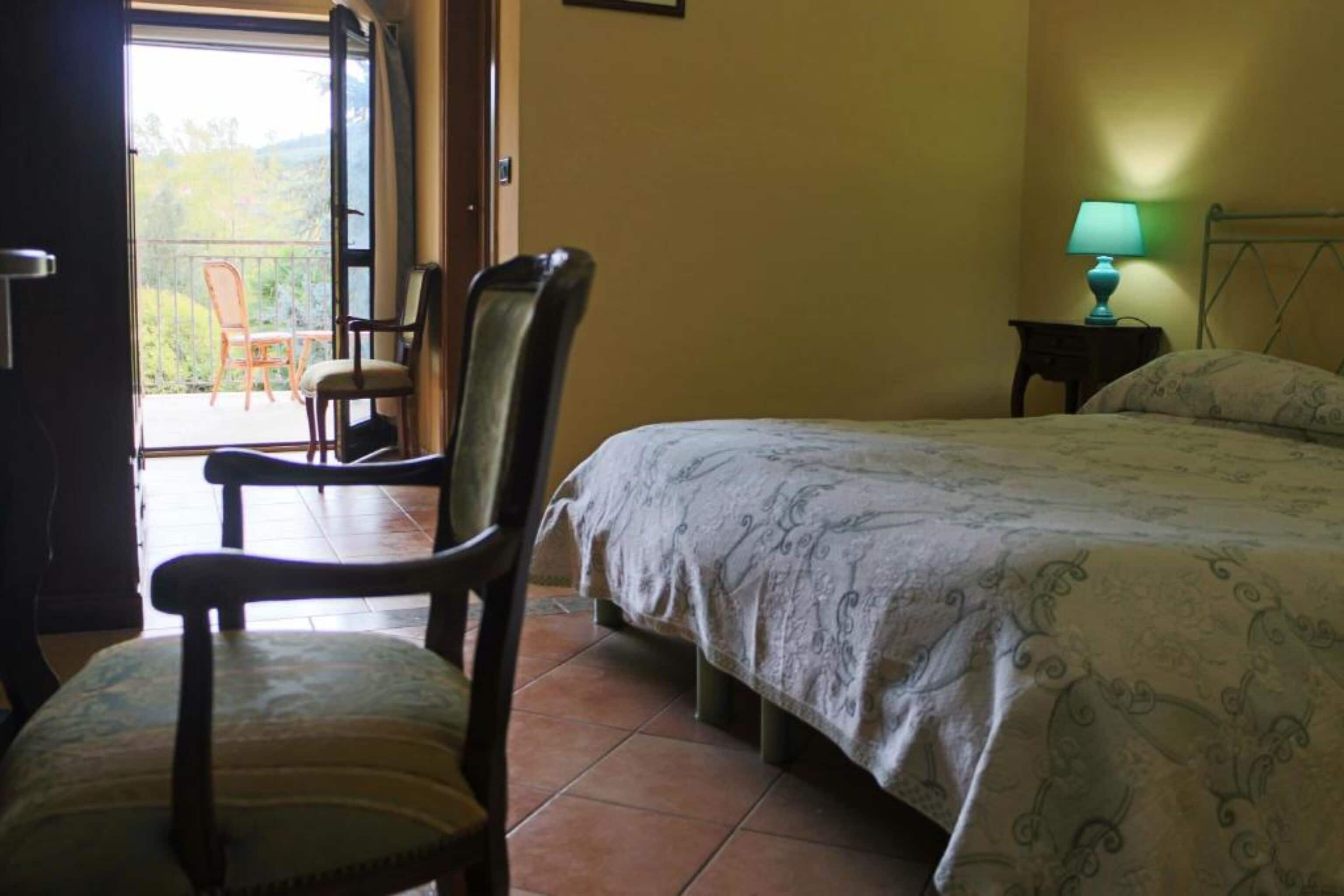 A bedroom with a bed, a chair, a nightstand with a lamp, and a view of a balcony with chairs.