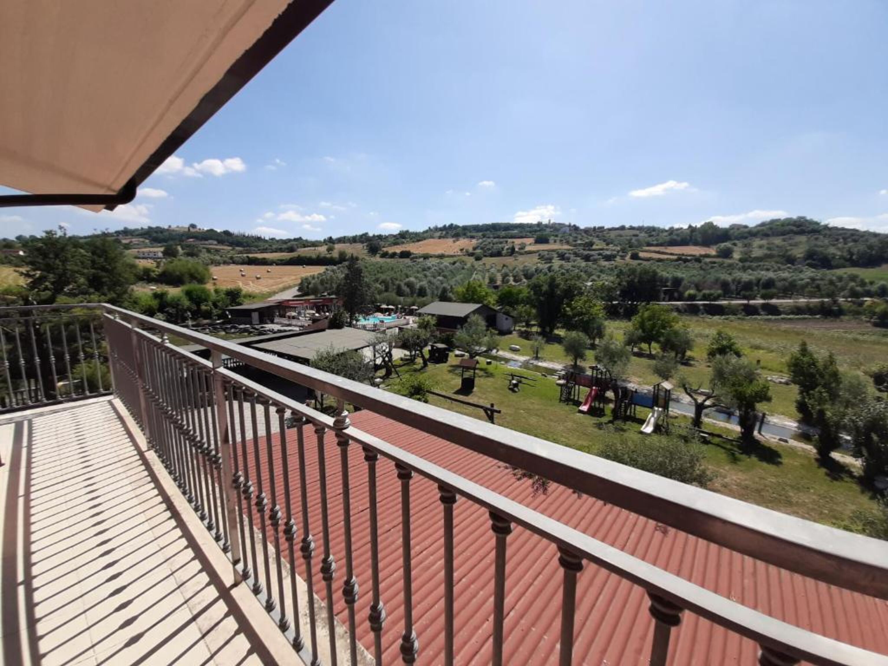 Un balcon doté d’un plancher rouge et d’une rambarde en métal, offrant une vue sur un paysage rural avec des champs et des arbres.