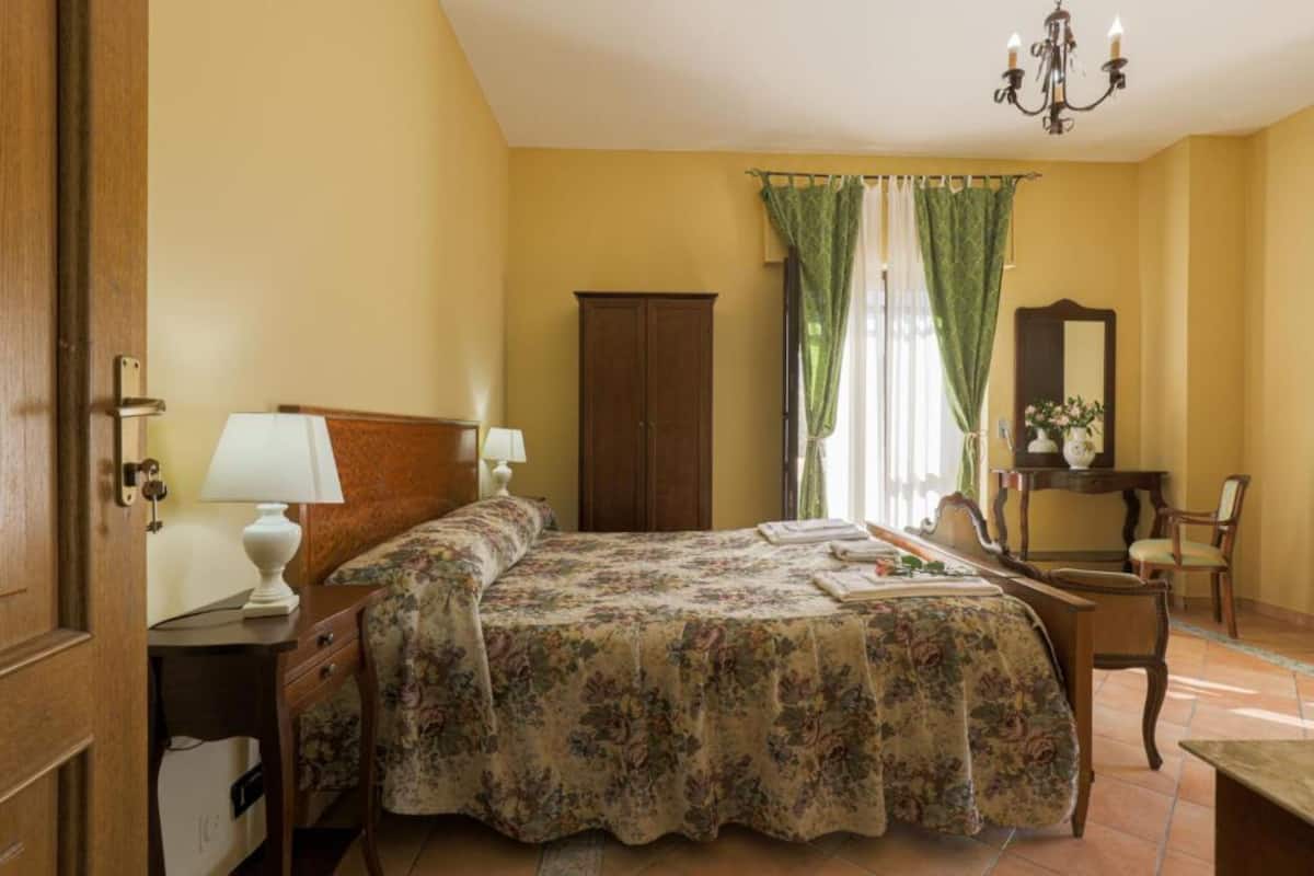 A bedroom with a bed, two bedside tables, a dresser, a chair, a desk, a mirror, and a chandelier.