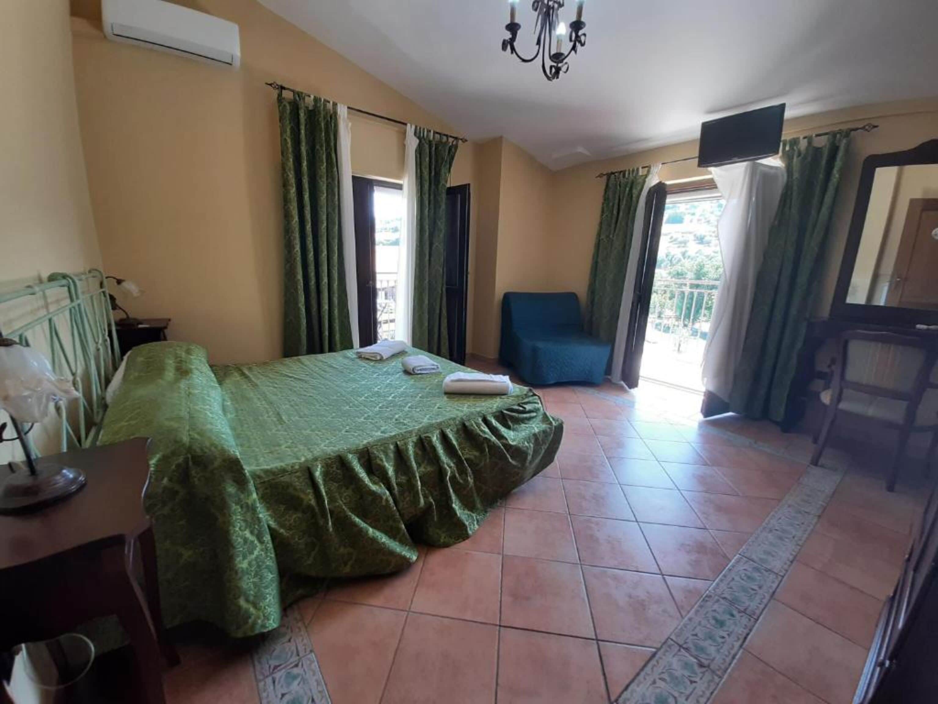 A hotel room with a bed, a chair, a television, and a balcony with curtains.