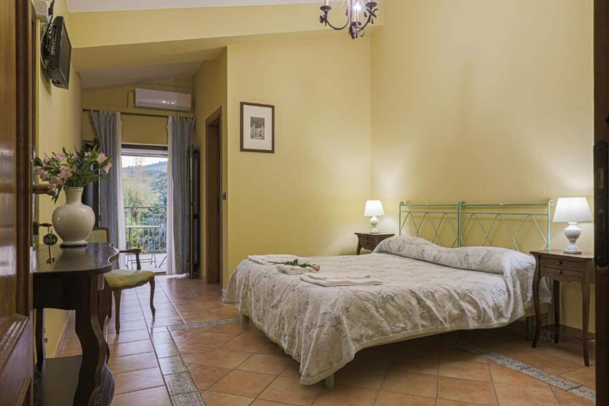 A bedroom with a bed, bedside lamps, a small table with a vase, and a view of the outdoors through a window.