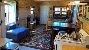 A cozy cabin interior with a sofa, a small table, a stove, and a bed with a window. - Secluded Off-Grid Cabin, Upper Peninsula Getaway on 36 Private Acres (Bruce Crossing)