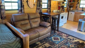 A cozy living room with a leather sofa, a dining area with a kitchen in the background, and a large window with curtains. - Secluded Off-Grid Cabin, Upper Peninsula Getaway on 36 Private Acres (Bruce Crossing)