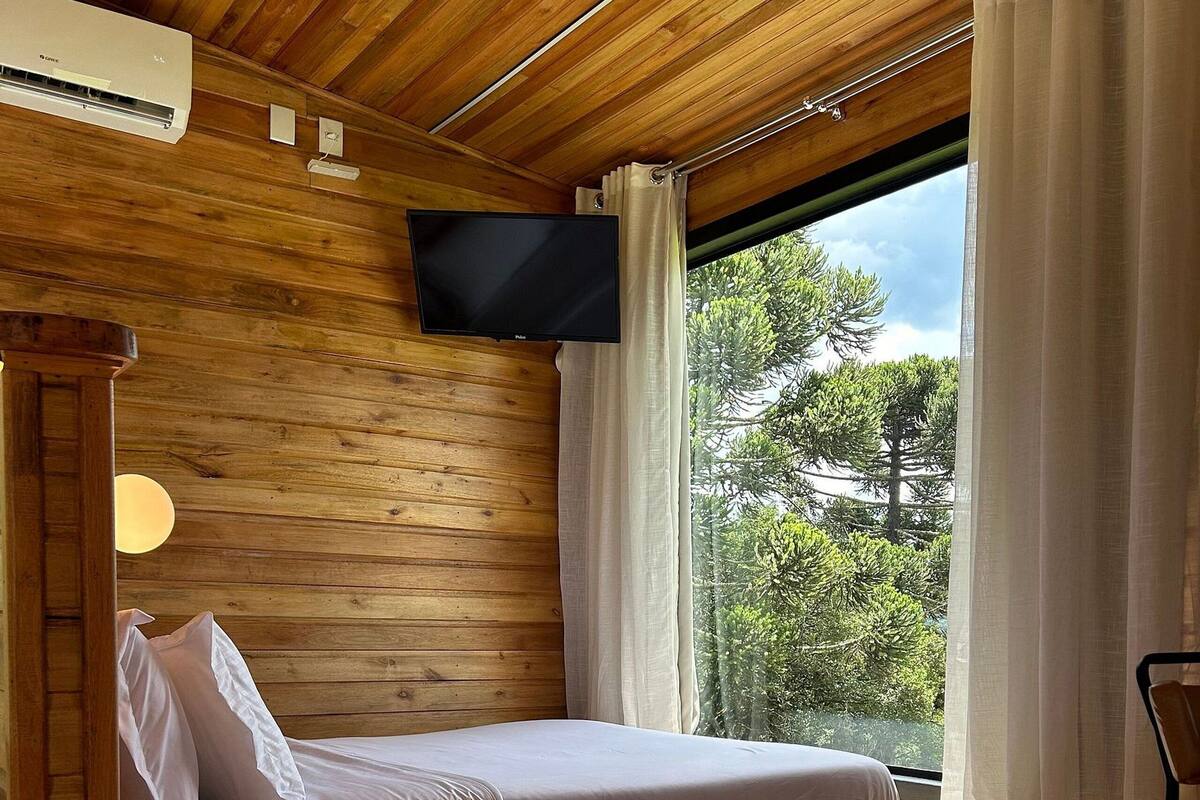 A wooden cabin interior with a bed, a mounted TV, and a window with curtains.