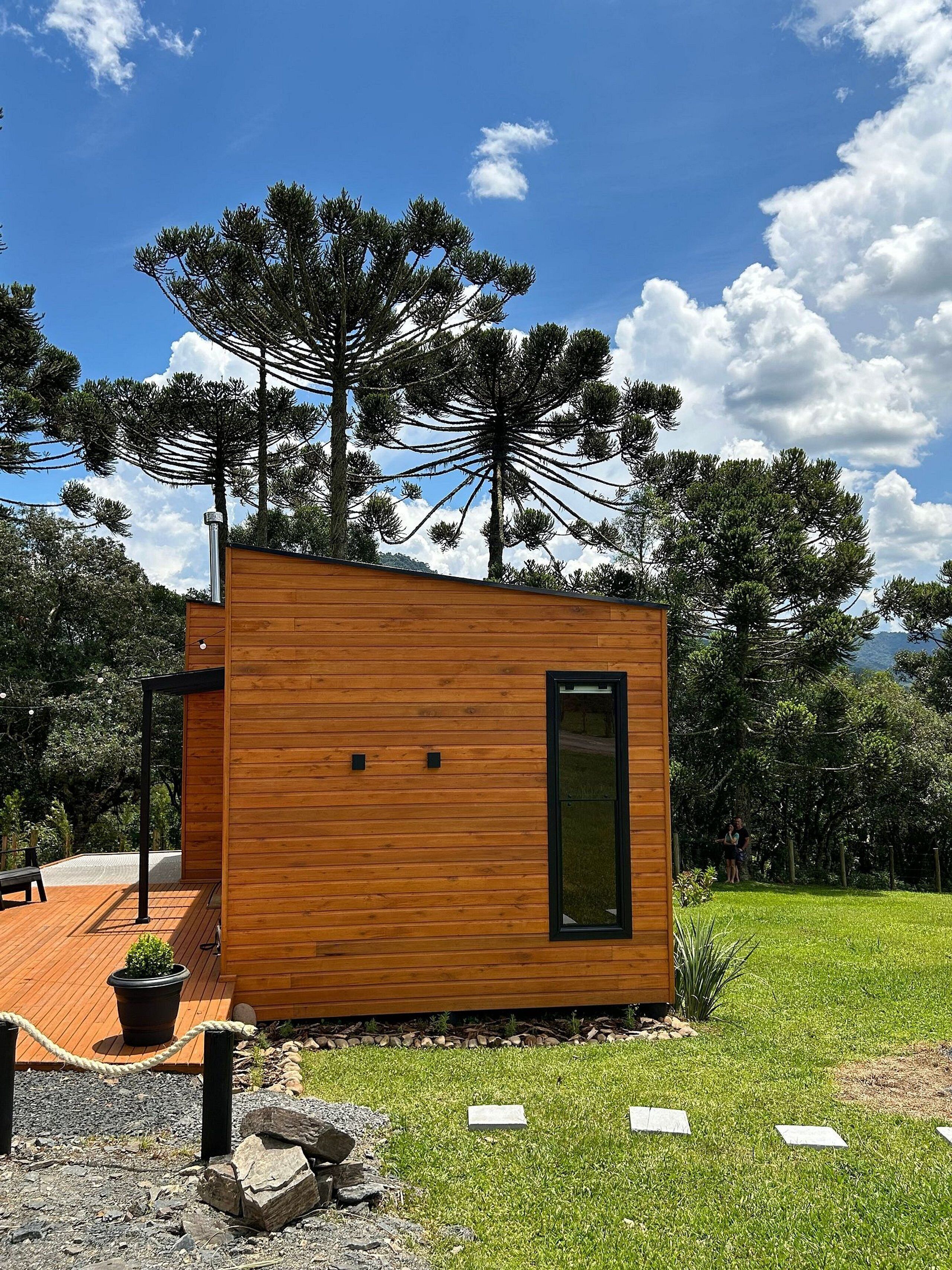 Une cabane en bois moderne, dotée d’une grande fenêtre et entourée d’une zone herbeuse et d’arbres.