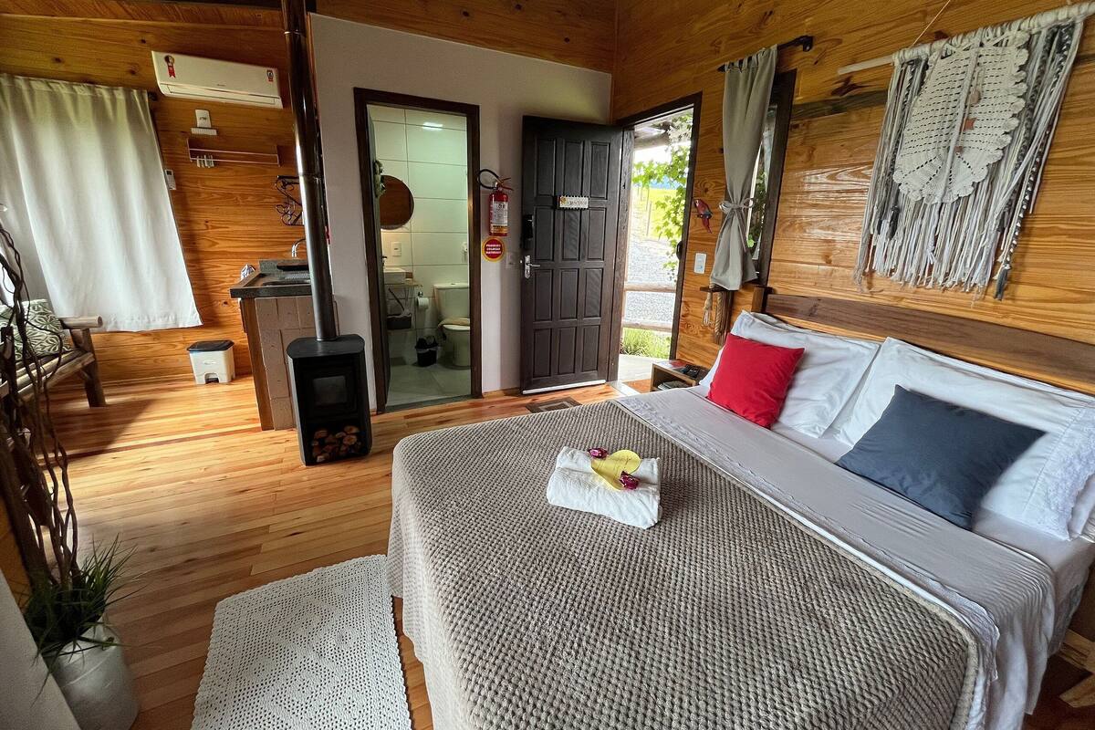 A cozy bedroom with a bed, a small bedside table, a window with curtains, and a wooden ceiling.
