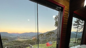 A modern bathroom with a large bathtub, a wooden ceiling, and a view of a rural landscape through large windows. - Chalés Solar da Montanha Urubici (Urubici)