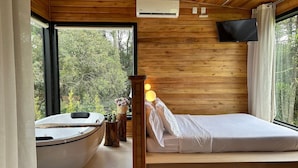A bedroom with a wooden ceiling, a large window, a bed, a bathtub, and a wall-mounted TV. - Chalés Solar da Montanha Urubici (Urubici)