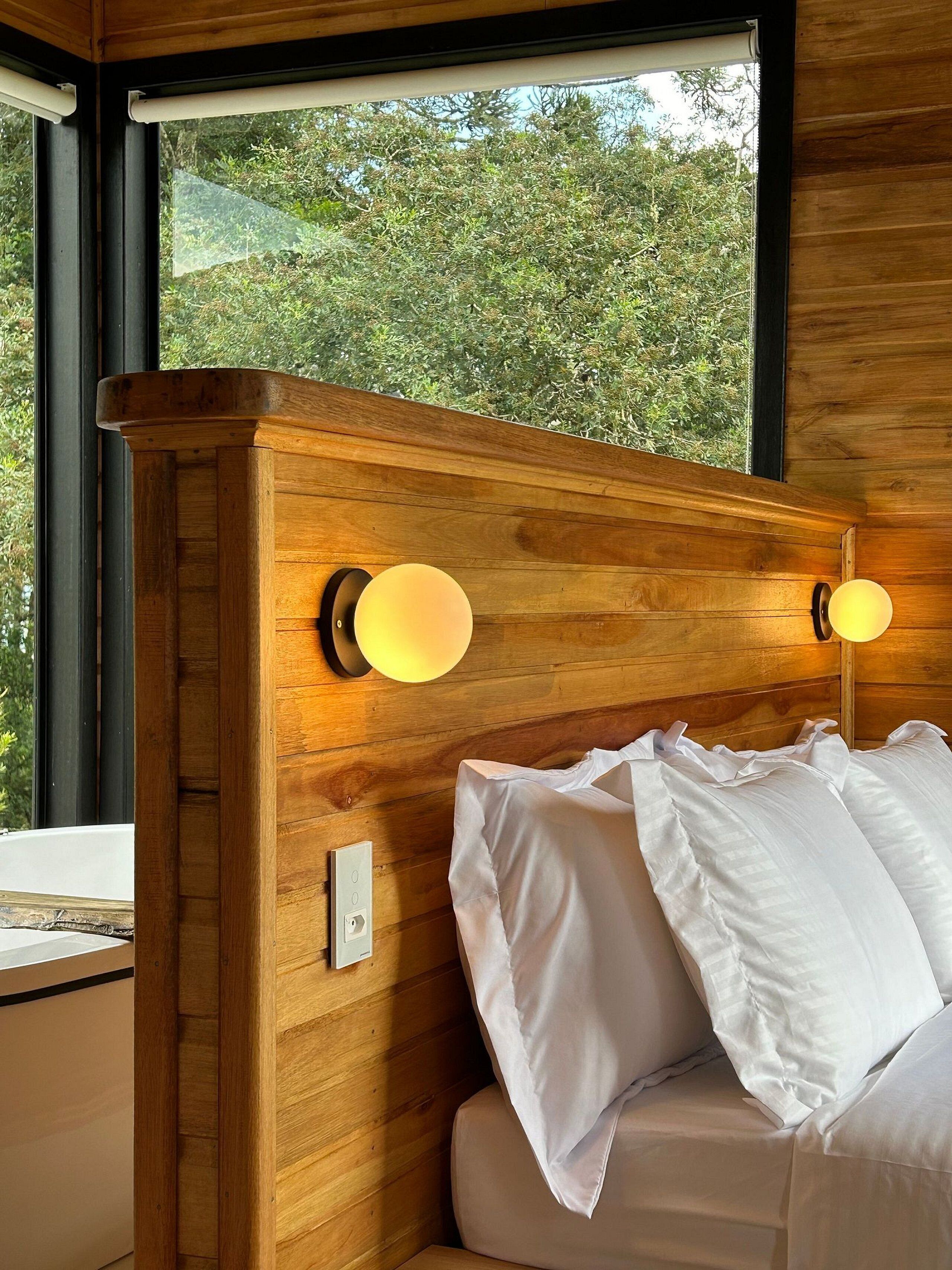 Cabecera de madera con dos lámparas de pared, cama con ropa de cama blanca y ventana con vistas a árboles.