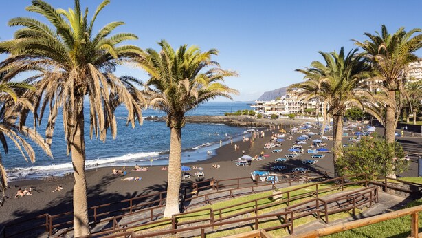 A beach with palm trees, a sandy shore, and a view of a resort area with buildings and parked cars. - Playa La Arena with pool and privat parking (Santiago del Teide)