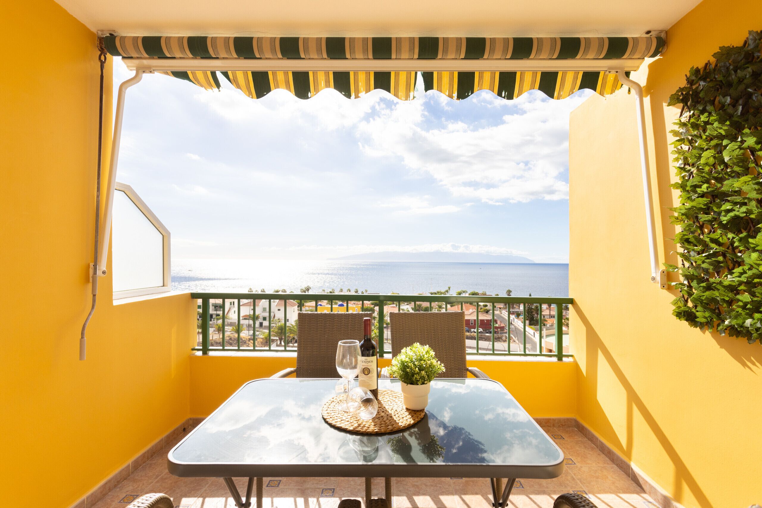 A balcony with a table, chairs, a bottle of wine, and a potted plant, overlooking a coastal view.