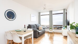 A modern living room with a dining area, a grey sofa, a ceiling fan, a large window with curtains, and a view of the city. - Playa La Arena with pool and privat parking (Santiago del Teide)