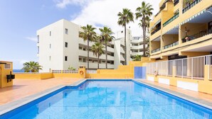 A swimming pool with a clear blue water surface, surrounded by a yellow building with balconies and palm trees. - Playa La Arena with pool and privat parking (Santiago del Teide)