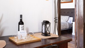 Una mesa de madera con una botella de vino, copas y un farol. Se ve una cama con una colcha estampada al fondo.
