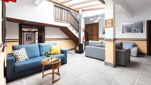 Amplia sala de estar con un sofá azul, un sofá gris y una pequeña mesa redonda con una planta. Una escalera con barandillas de madera conduce a un nivel superior.