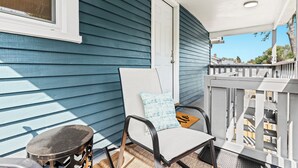 A balcony with a chair and a small stool, a white door, and a view of the neighborhood.