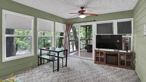 Una habitación con ventilador de techo, un televisor de pantalla plana sobre un soporte de madera, una mesa de comedor con sillas y ventanas amplias con cortinas.