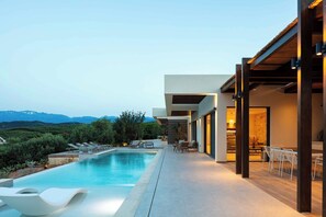 A modern outdoor pool area with lounge chairs and a dining area, overlooking a mountainous landscape. - Villa Aglaea (Platanias)