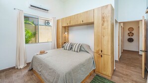 Ein Schlafzimmer mit Bett, Holzschrank, Fenster mit Vorhängen und einem durch die Tür erkennbaren kleinen Badezimmer.