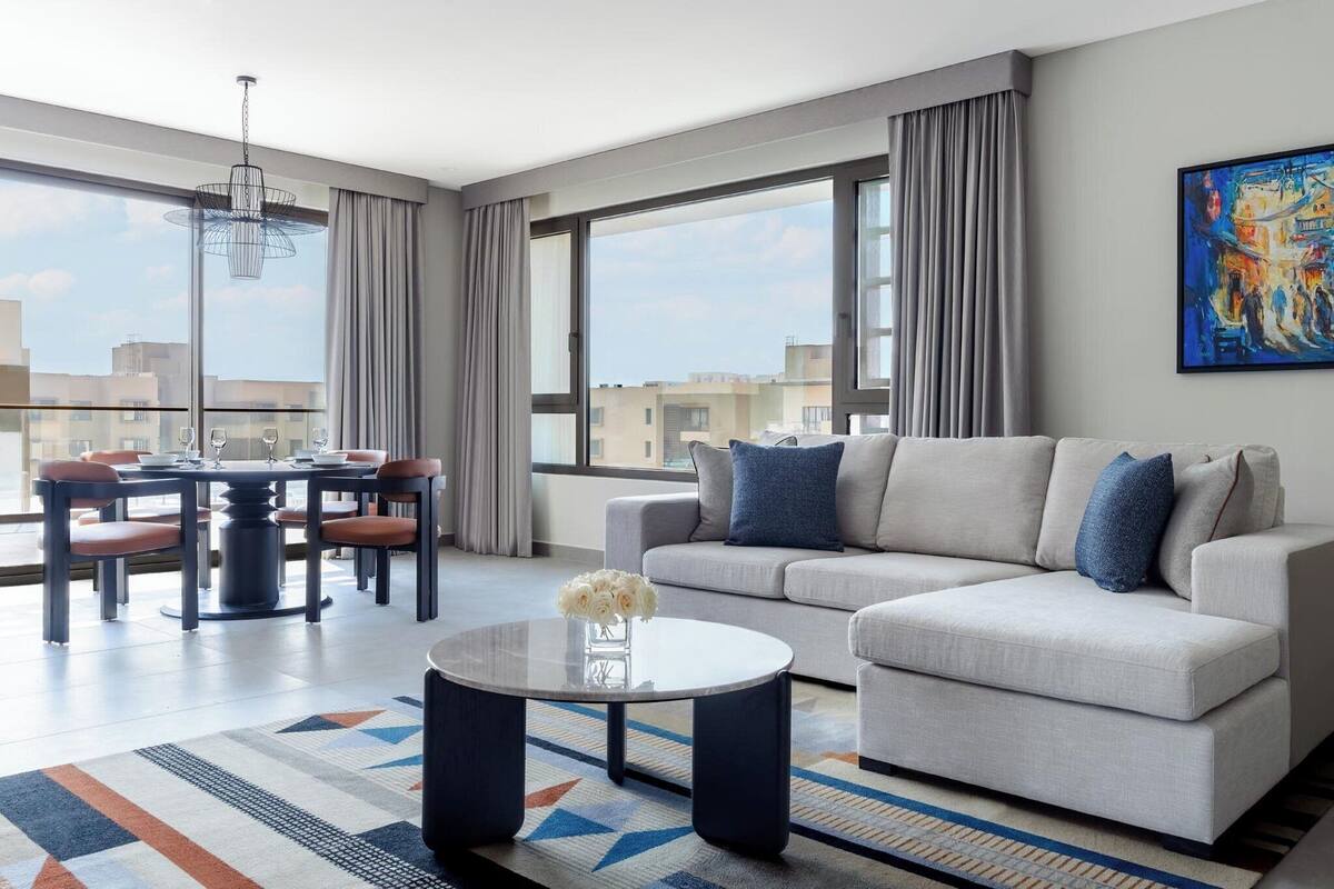 A modern living room with a sofa, a dining table with chairs, a coffee table with flowers, and large windows with curtains.