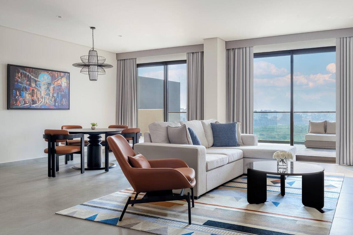 A modern living room with a sofa, armchairs, a dining table, and a large window with a city view.