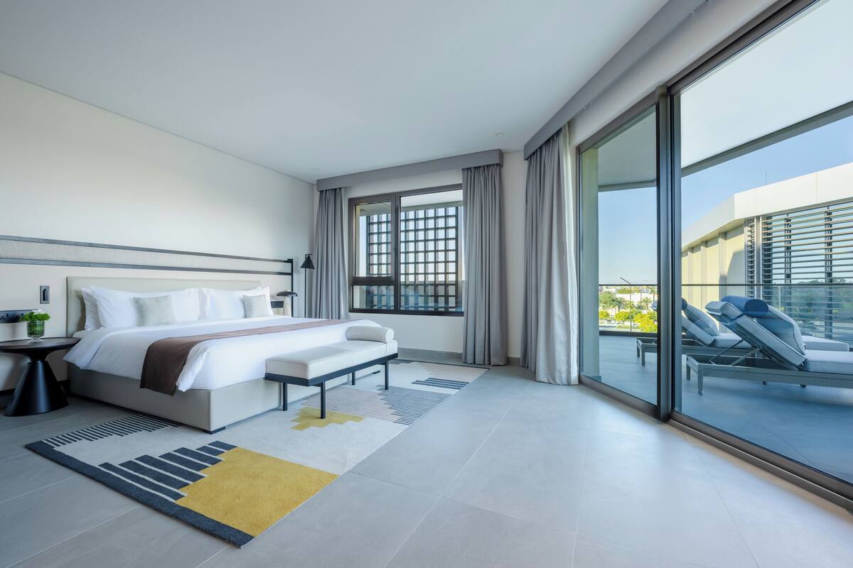 A modern hotel room with a large bed, a bedside table, a small desk, a chair, a large window with curtains, and a view of the outside balcony.