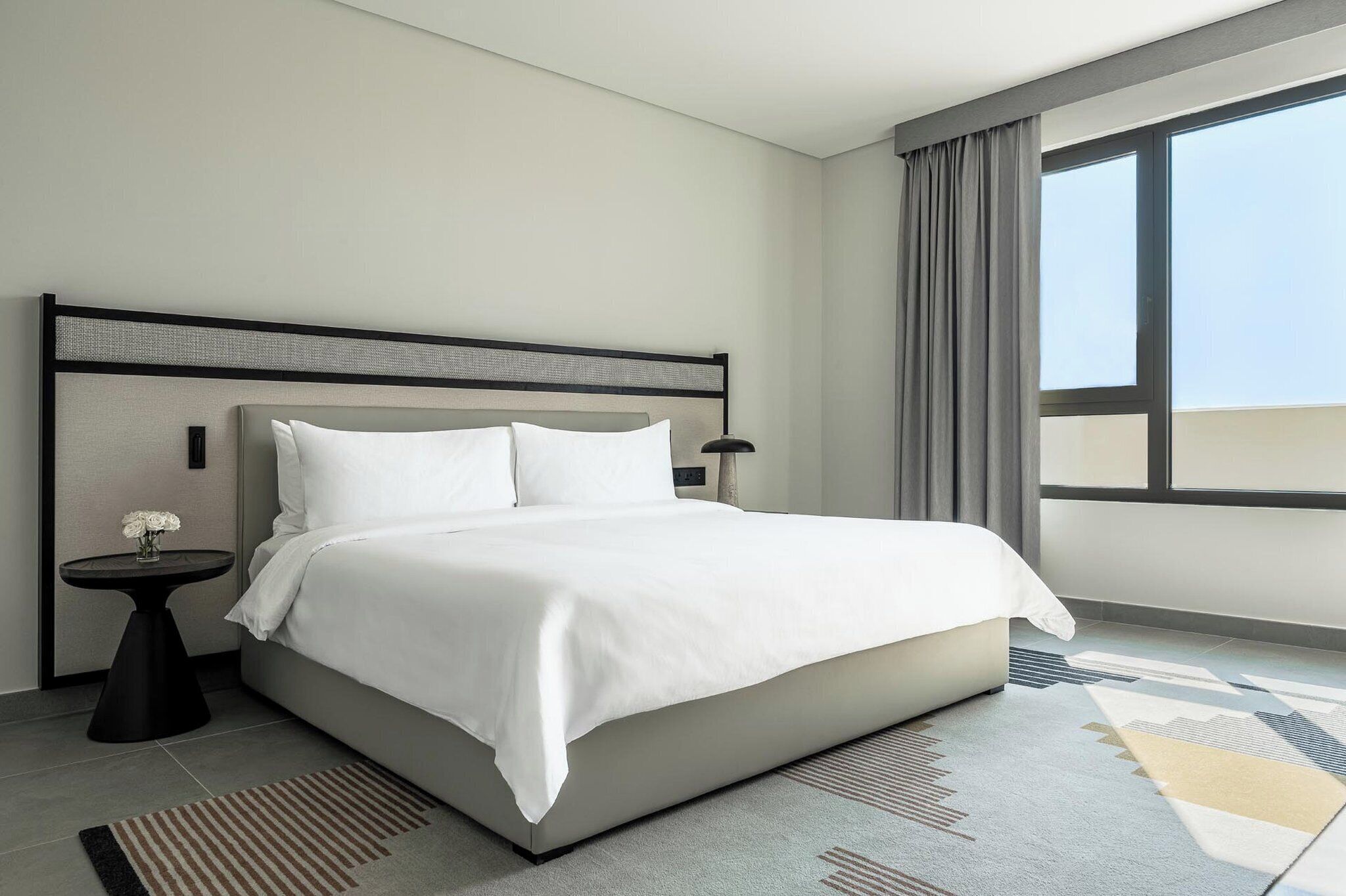 A modern hotel room with a large bed, a round side table with a vase of flowers, a lamp, and a view of the sky through a large window.