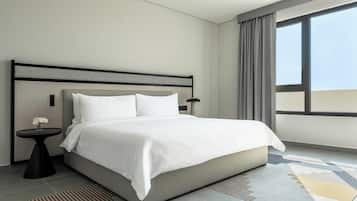 A modern hotel room with a large bed, a round side table with a vase of flowers, a lamp, and a view of the sky through a large window.