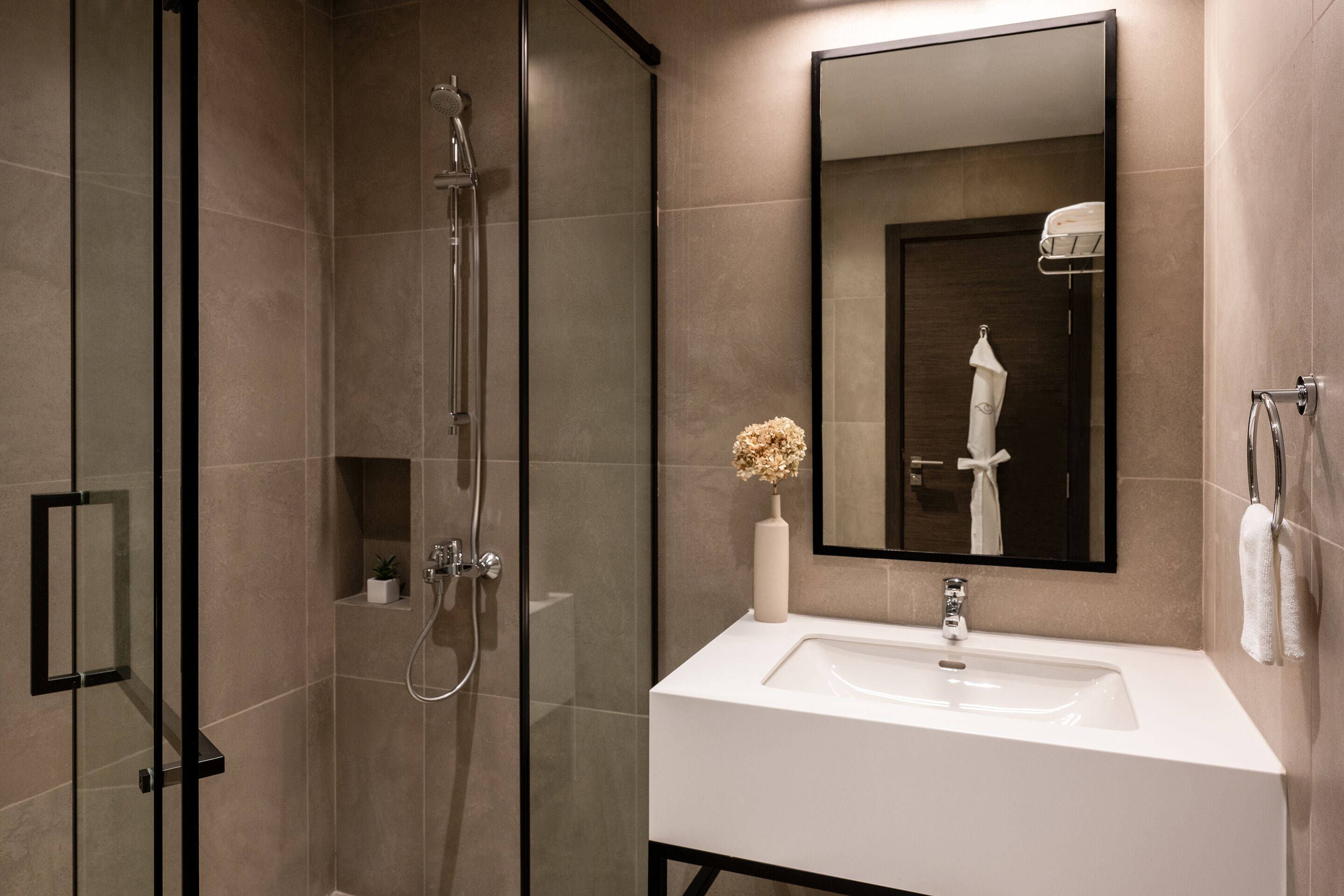 Un baño moderno con ducha envuelta en vidrio, un lavamanos blanco, un espejo grande y un florero con flores secas.