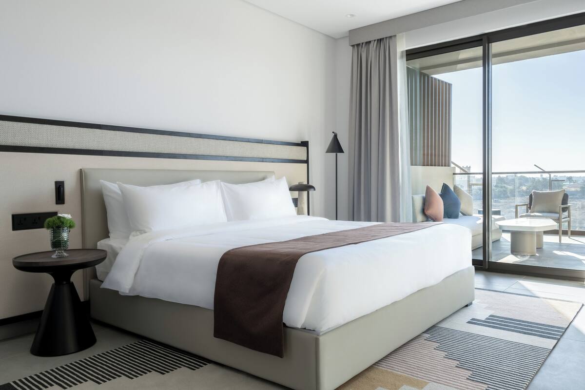 A modern hotel room with a large bed, a small side table, and a balcony with a view.