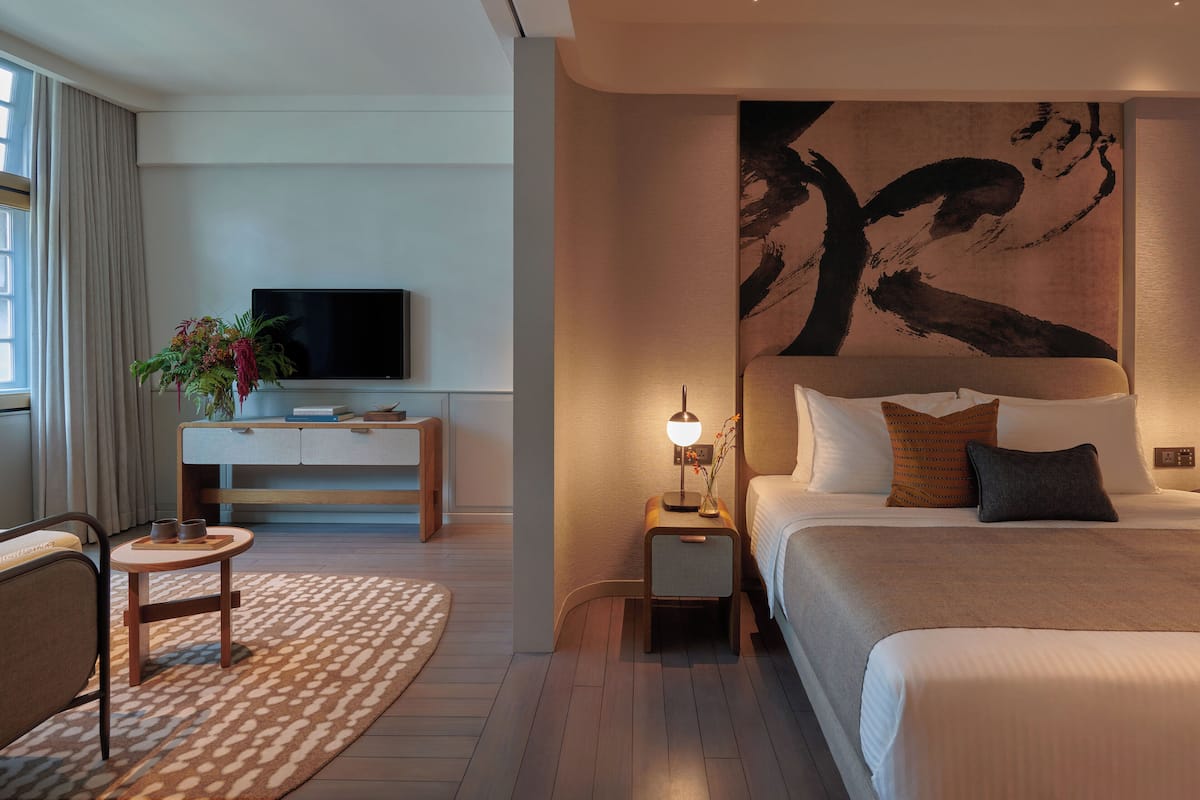 A modern hotel room with a large bed, a TV on a wooden console, a small side table with a lamp, and a large abstract painting on the wall.