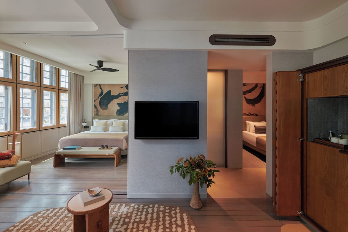A modern hotel room with a large bed, a flat-screen TV mounted on the wall, a wooden wardrobe, and a large window offering a view of the city.