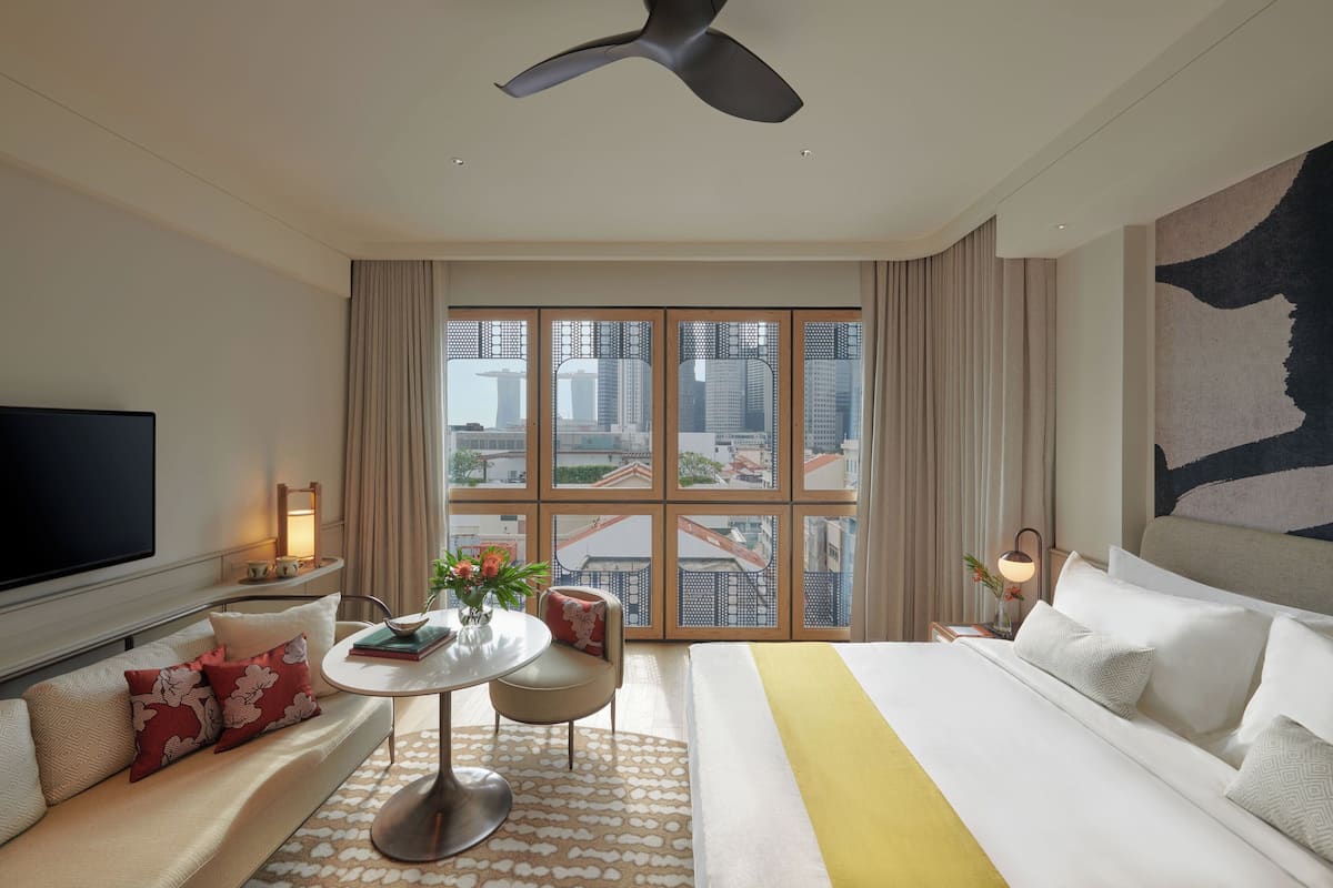 A hotel room with a large bed, a TV, a dining table with chairs, a small table with a vase of flowers, and a view of the city through large windows.