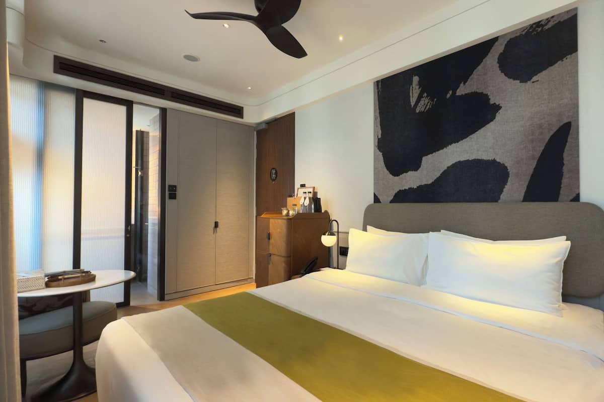 A hotel room with a large bed, a desk, a chair, a wardrobe, and a wall with a large abstract painting.