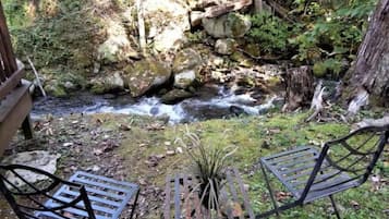 A natural outdoor setting with a small stream, two metal chairs, and a wooden bench.