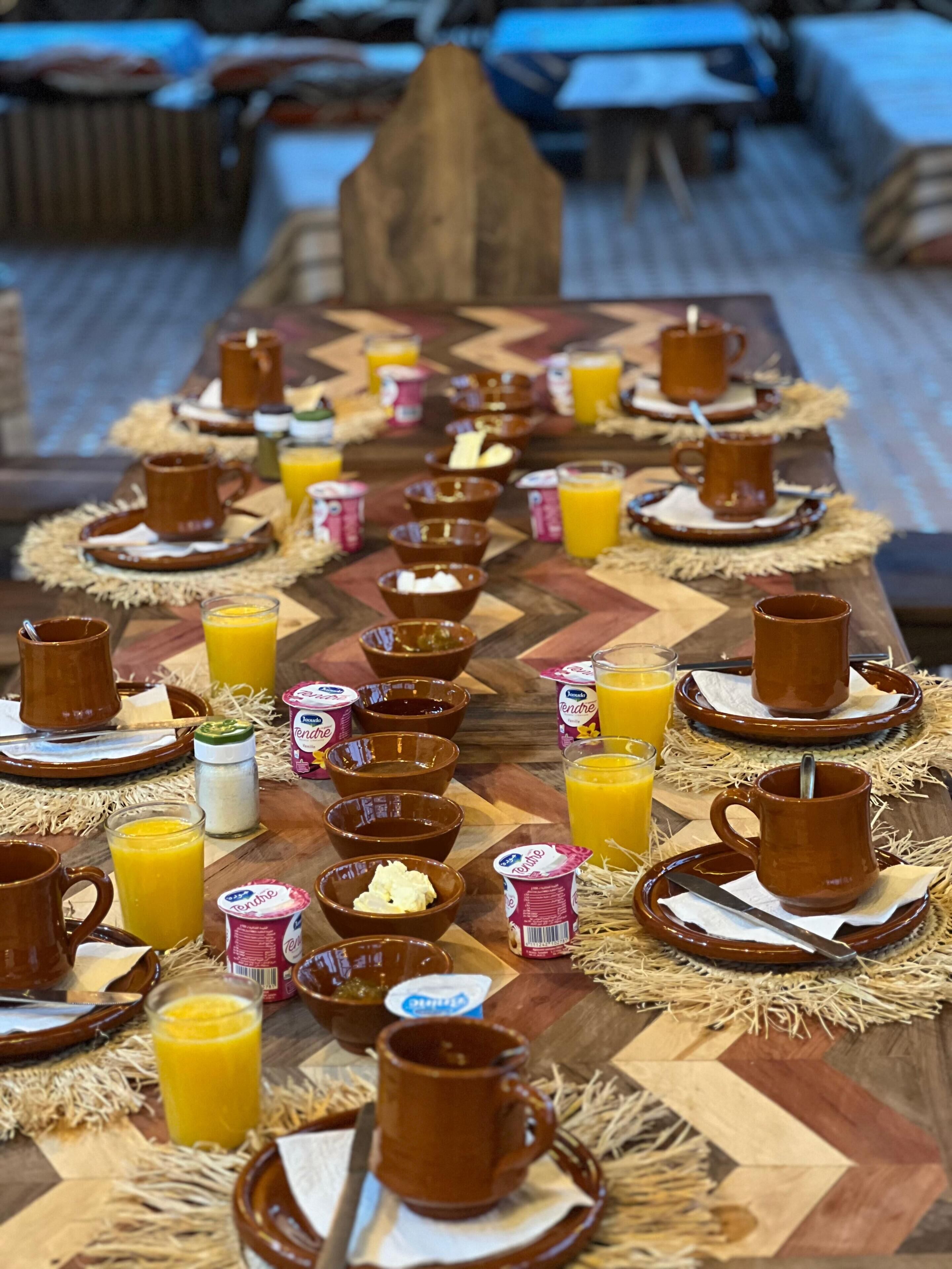 A long dining table set with multiple mugs, glasses of orange juice, and bowls of food, likely a breakfast buffet.