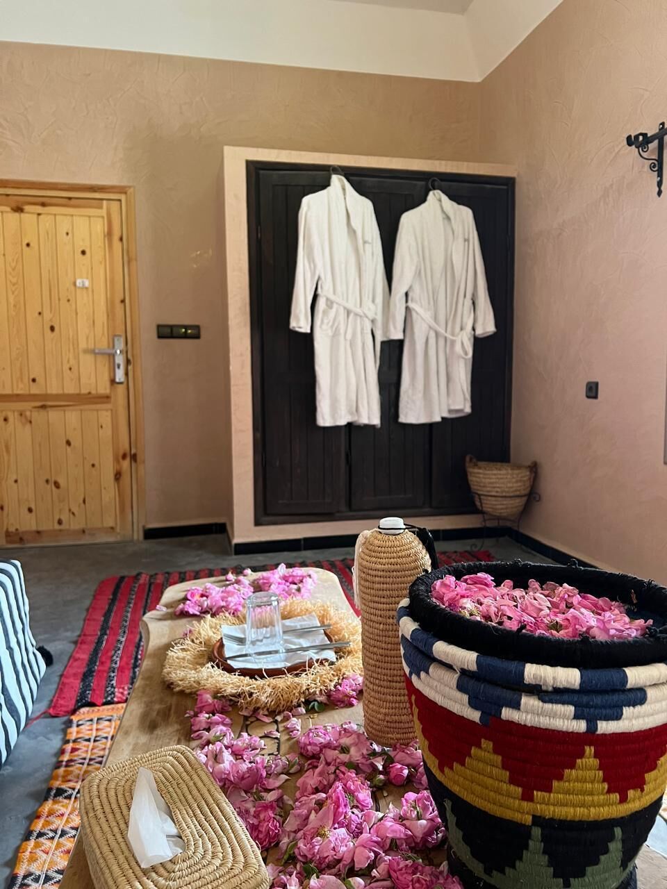 A room with a wooden door, a black-framed mirror, a woven basket with pink flowers, and a woven mat on the floor.