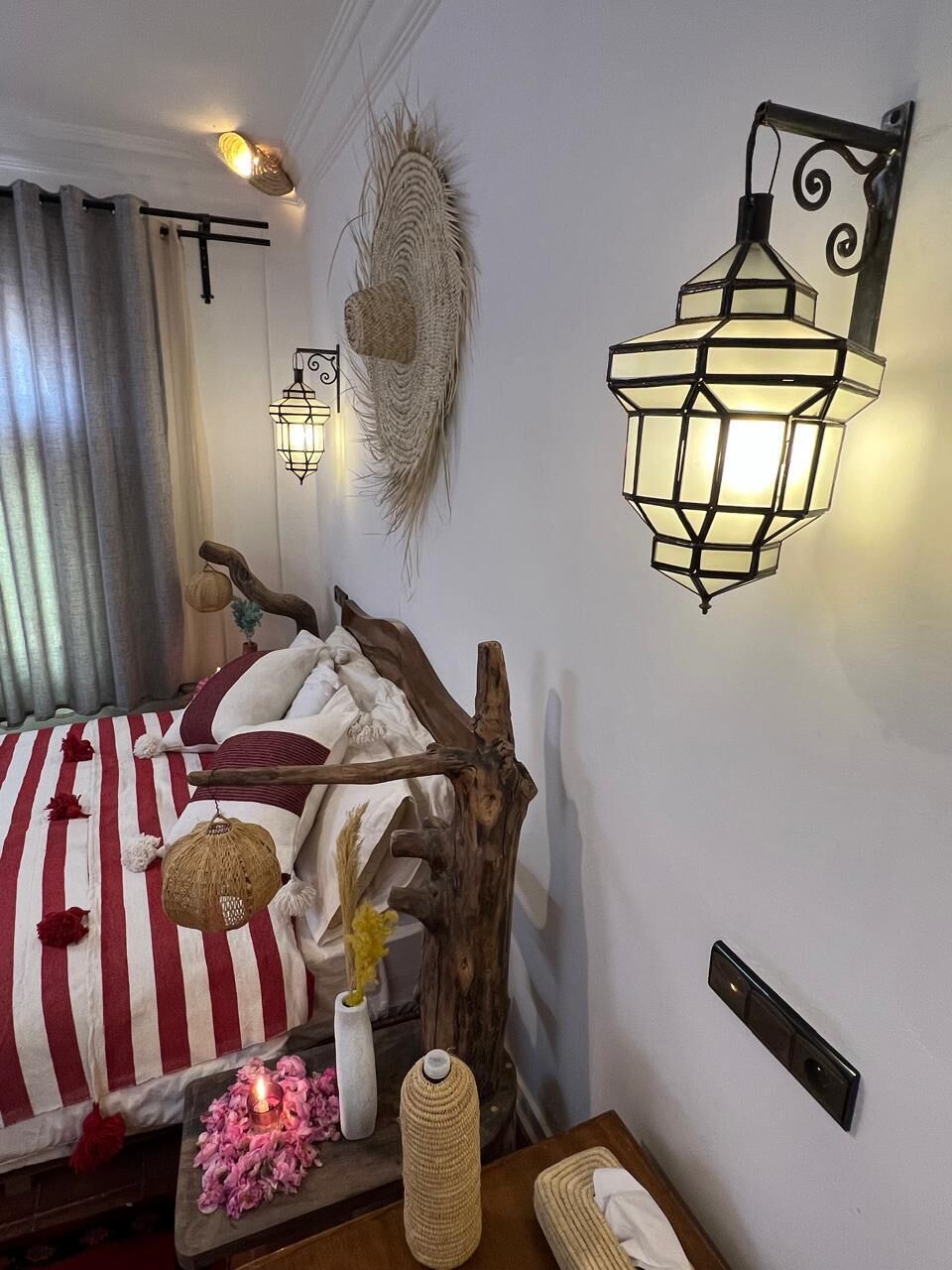 Um quarto com uma cama, uma luminária na parede, um objeto decorativo na parede e uma mesa de cabeceira com uma vela e um vaso.
