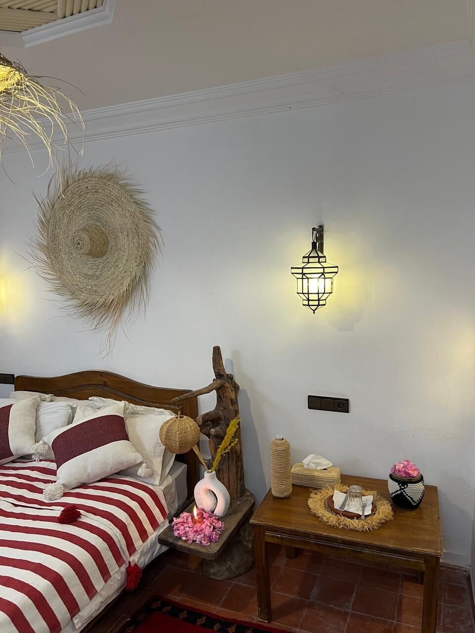 Een slaapkamer met een bed, een wandlamp en een klein houten tafeltje met decoratieve objecten.