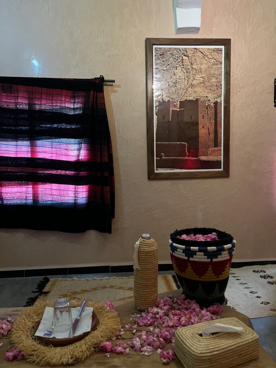 A room with a framed photograph on the wall, a woven rug, a small table with a glass and a fork, and a decorative basket.
