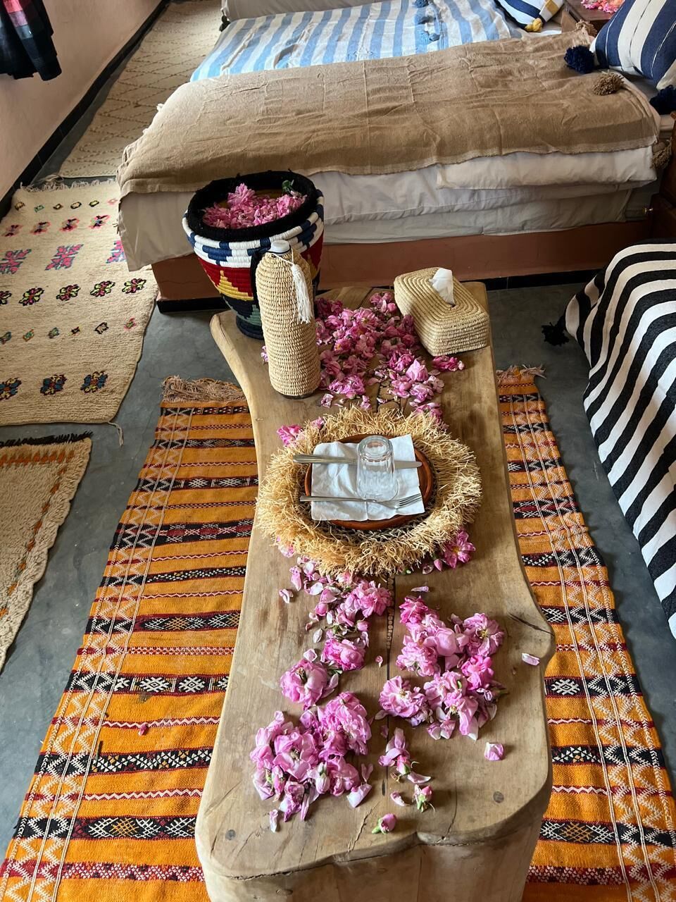A room with a bed, a wooden table with a glass, a woven basket, and a patterned rug.