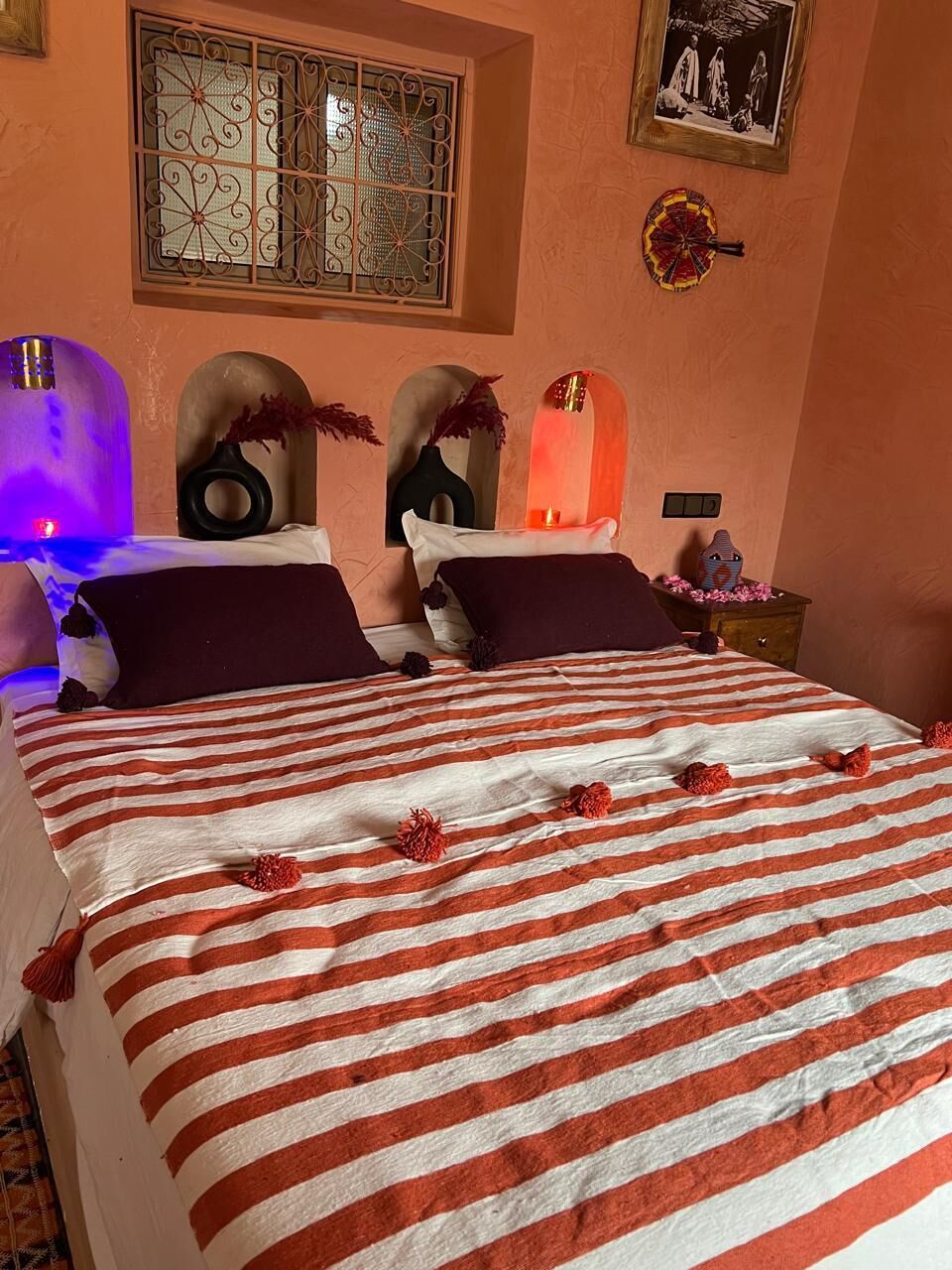 A bed with a striped bedspread, two pillows, a vase with dried flowers, a wall-mounted picture, and a decorative wall piece.