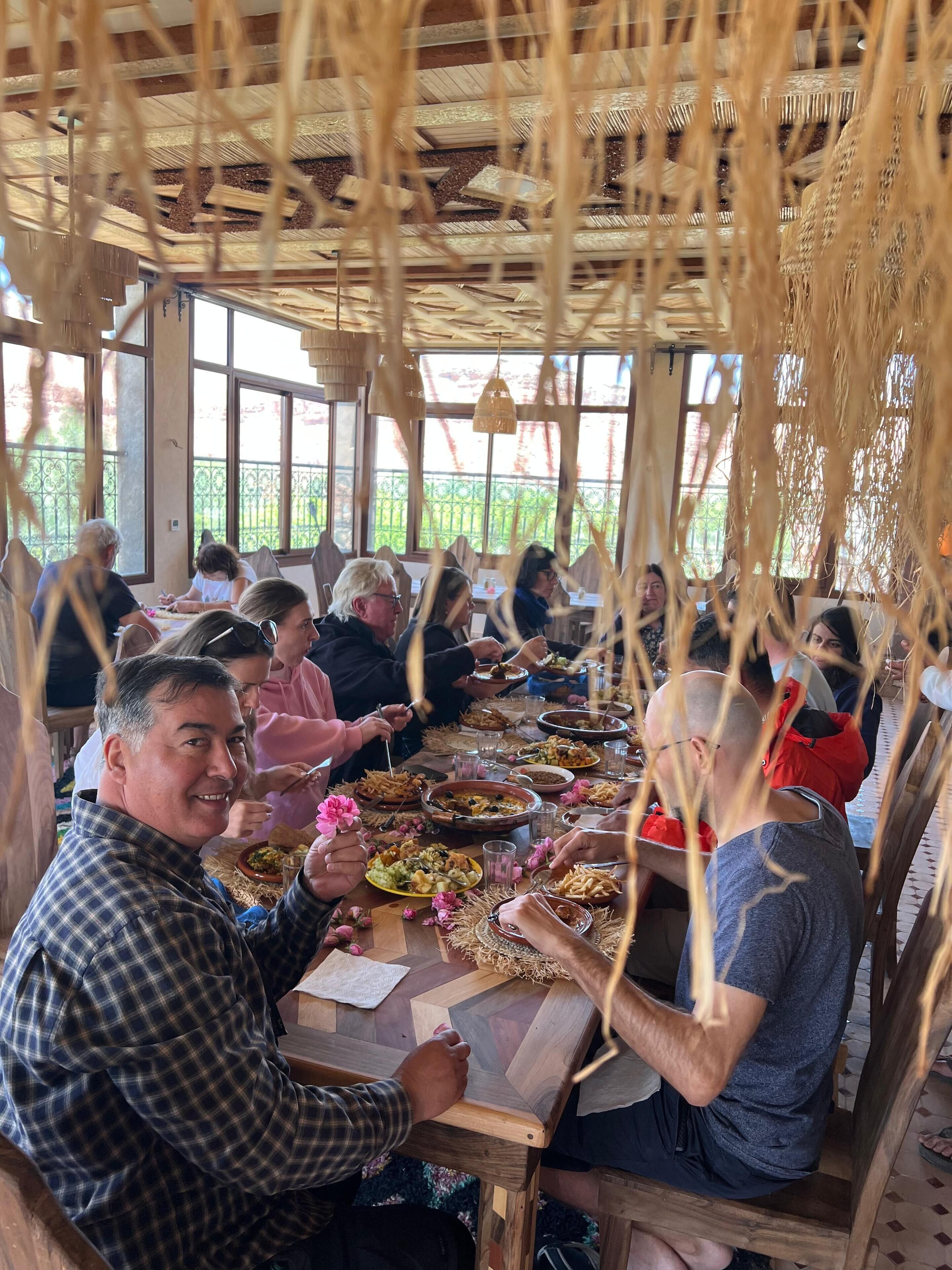 A group of people dining at a wooden table with a rustic ceiling, large windows, and a view of greenery outside.