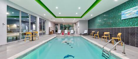 A modern indoor pool with clear blue water, surrounded by yellow chairs and a green accent wall.
