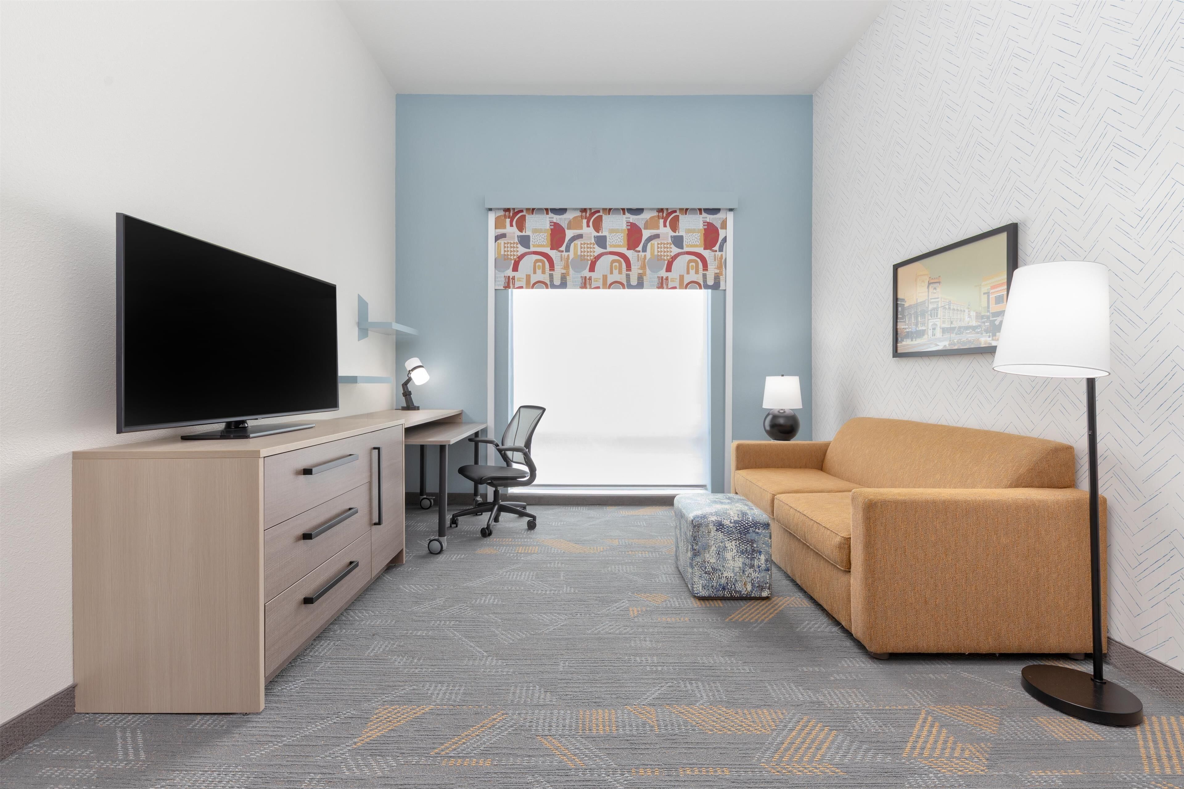 A hotel room with a TV on a wooden cabinet, a desk with a chair, a sofa, a patterned rug, and a window with a decorative valance.