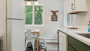 Une cuisine avec des meubles blancs, une table à manger en bois avec des chaises, une fenêtre donnant sur un espace verdoyant et un luminaire fixé au mur.