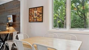 Un coin repas avec une table en bois et des chaises blanches, une œuvre d’art encadrée au mur et une fenêtre donnant sur des arbres.