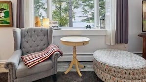 Une pièce avec un fauteuil gris, une table ronde et un pouf à motifs. Une fenêtre donne sur des arbres et on peut y voir une enseigne sur laquelle est écrit « THE CABIN ».