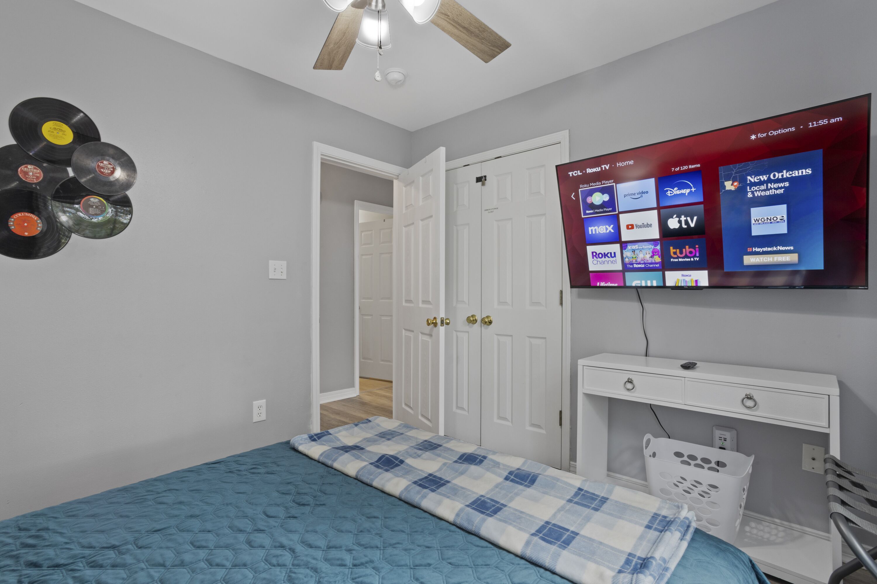 Une chambre à coucher avec un lit, un téléviseur fixé au mur qui diffuse différentes chaînes, et un ventilateur de plafond.