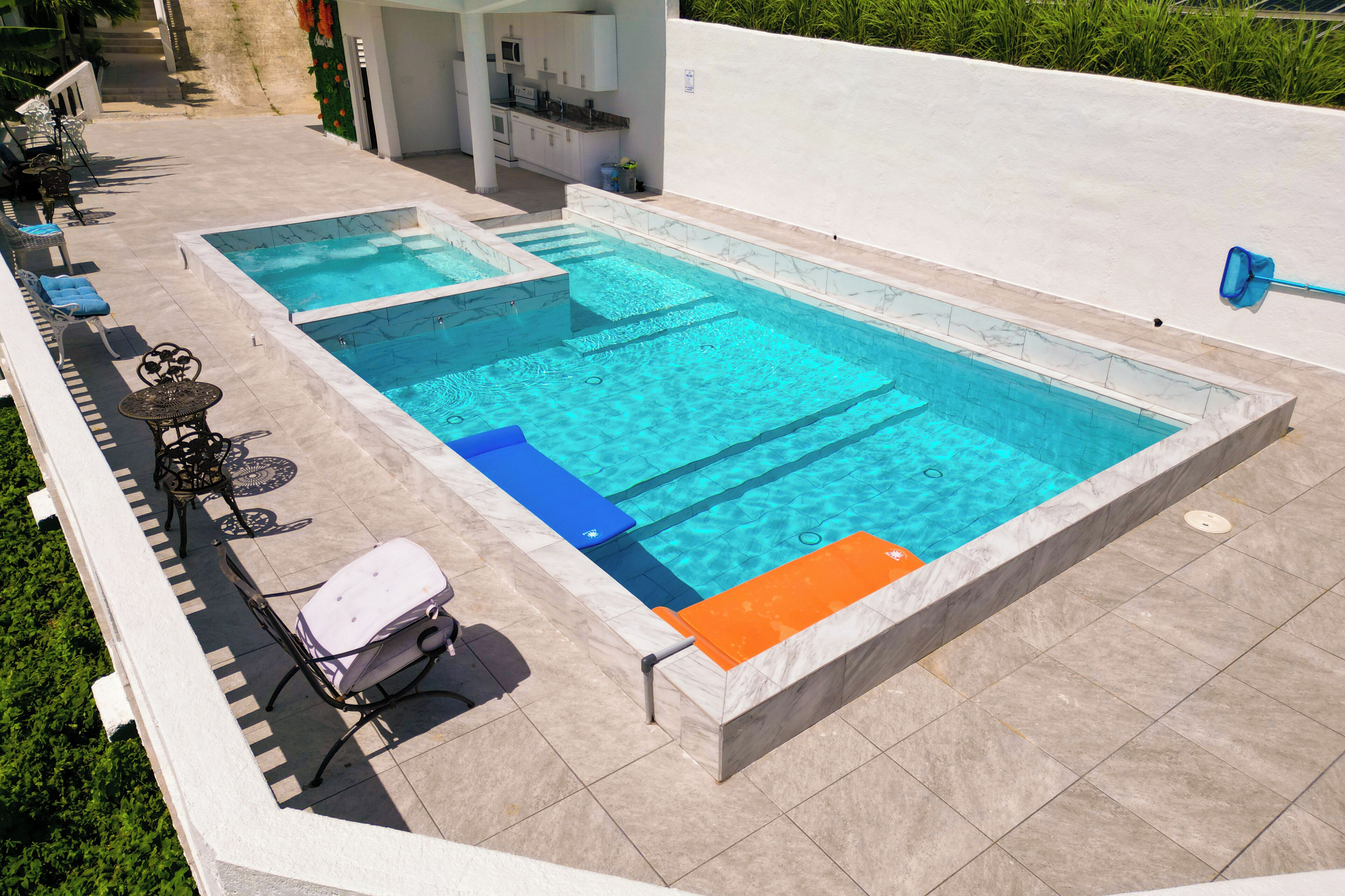A rectangular swimming pool with two smaller pools, surrounded by a tiled deck with lounge chairs and a table.